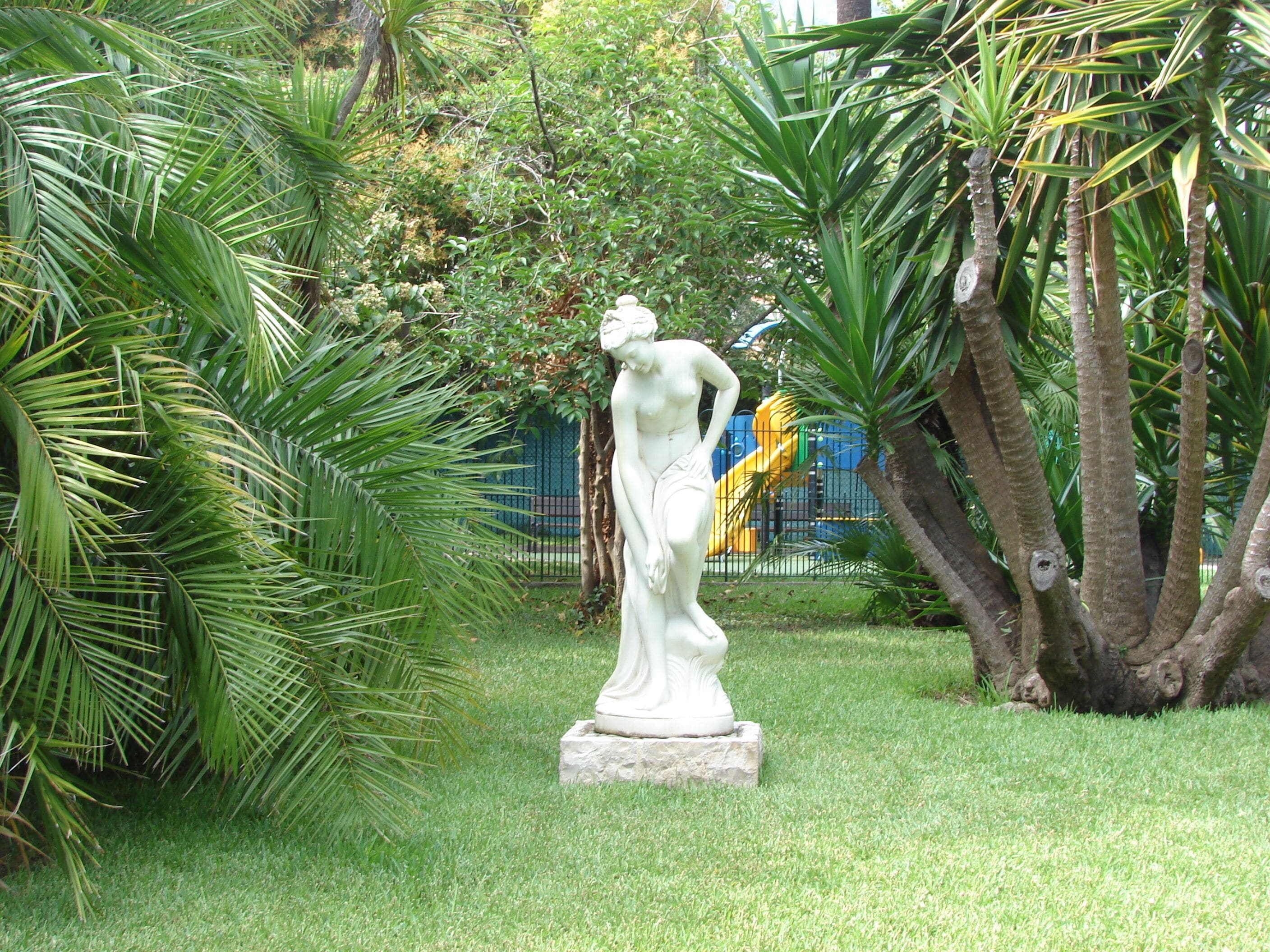 Una estatua en un jardín con palmeras y un parque infantil al fondo.
