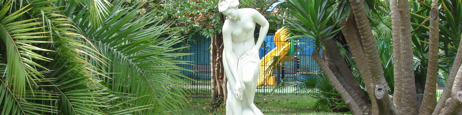 Una estatua en un jardín con palmeras y un parque infantil al fondo.