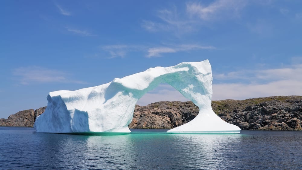 A large iceberg with a distinct arch shape floating in calm waters, surrounded by rocky terrain and a clear blue sky.