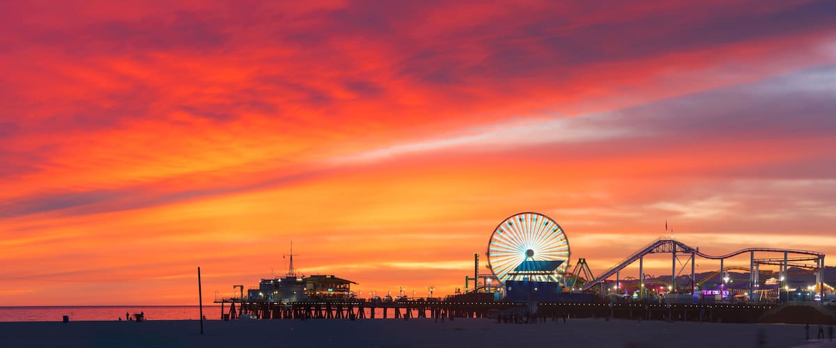 A pier with a large Ferris wheel and amusement rides against a sunset sky.