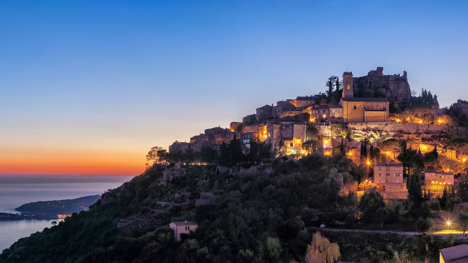 A historic fortified town perched on a hill overlooking a calm sea at dusk.