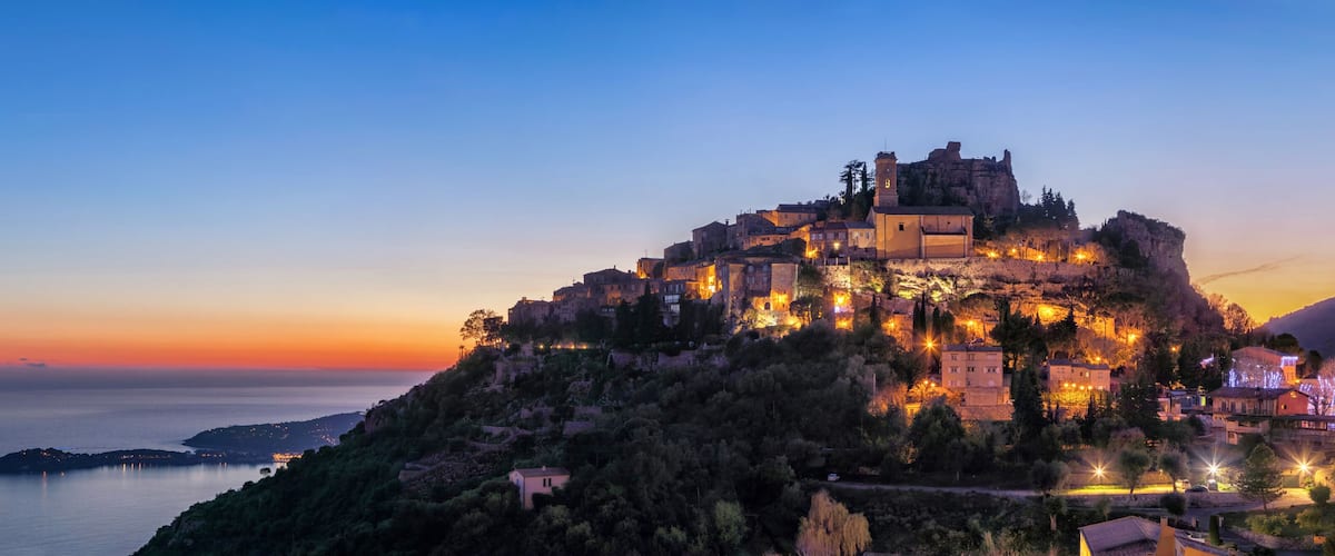 A historic fortified town perched on a hill overlooking a calm sea at dusk.