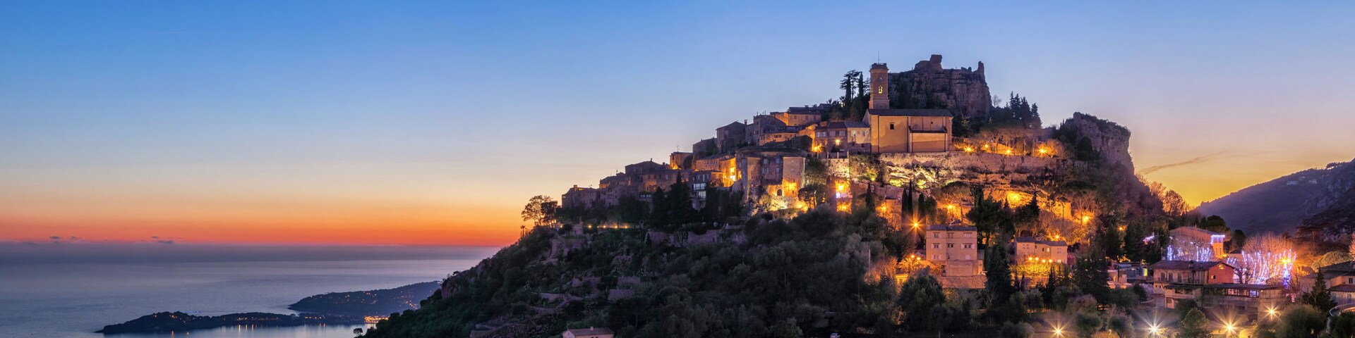 A historic fortified town perched on a hill overlooking a calm sea at dusk.