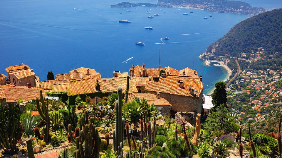 A coastal town with terracotta-roofed buildings, a cactus garden, and boats in the sea.