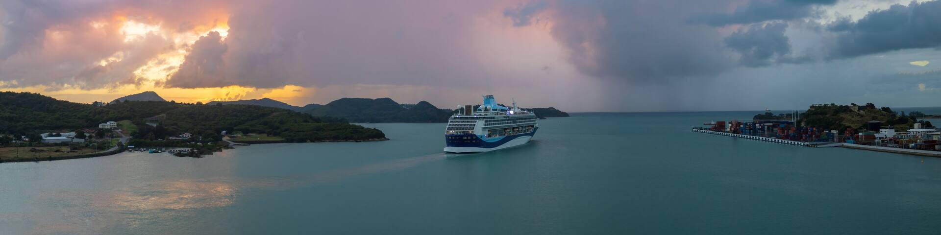 A cruise ship sailing in a calm bay with a backdrop of mountains and a dramatic sky.