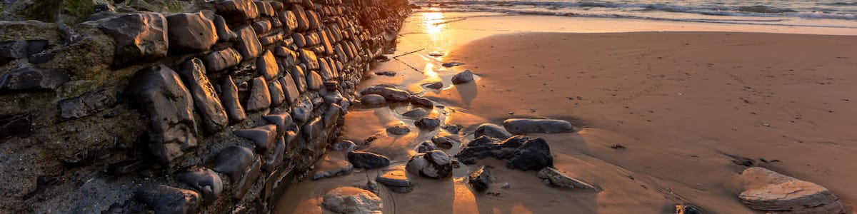 A sunset over a rocky beach with waves, a stone wall, and a clear sky.