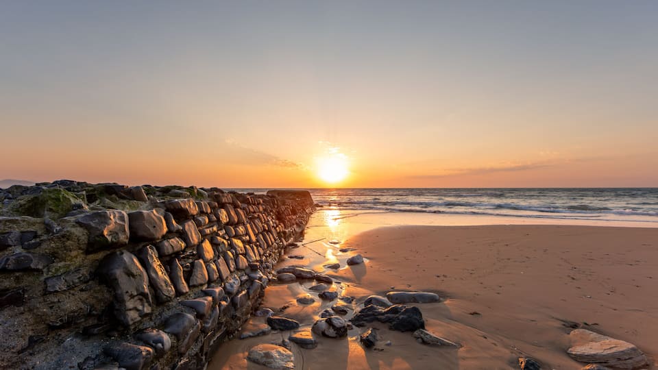A sunset over a rocky beach with waves, a stone wall, and a clear sky.