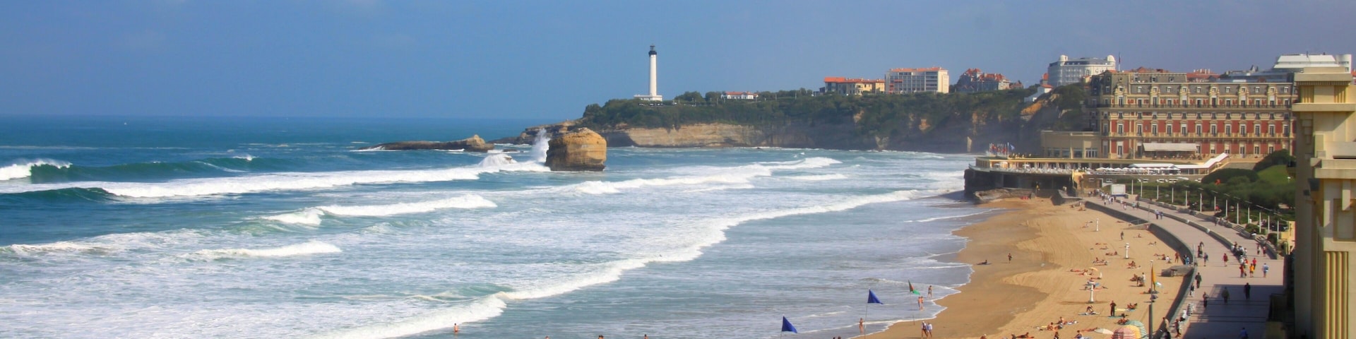 A coastal cityscape with a beach, waves, a lighthouse, and buildings.
