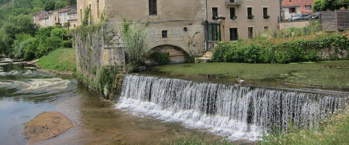 Un edificio histórico de piedra con una cascada frente a él, rodeado de exuberante vegetación y un río.