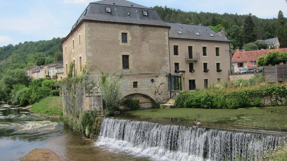 A historic stone building with a waterfall in front, surrounded by lush greenery and a river.
