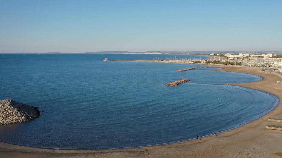 A coastal beach with calm waters, a rocky outcrop, and a row of buildings along the shore.