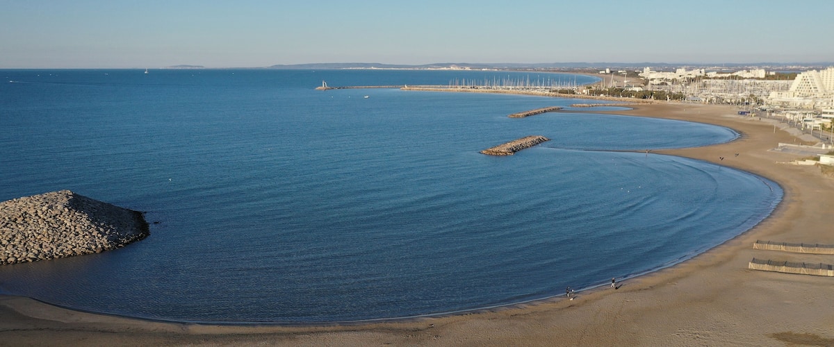 A coastal beach with calm waters, a rocky outcrop, and a row of buildings along the shore.
