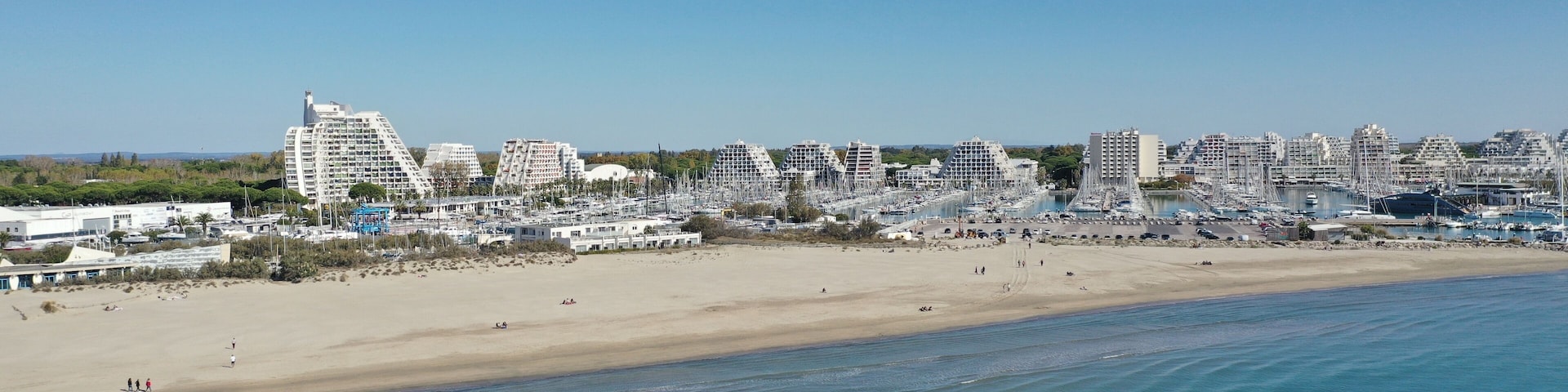 Un paisaje costero con una playa arenosa, un puerto deportivo lleno de barcos y modernos edificios altos.