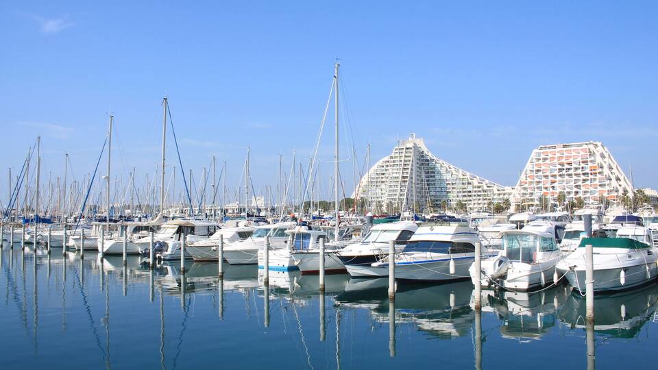 A marina with numerous boats docked, a clear blue sky, and distinctive modern buildings in the background.