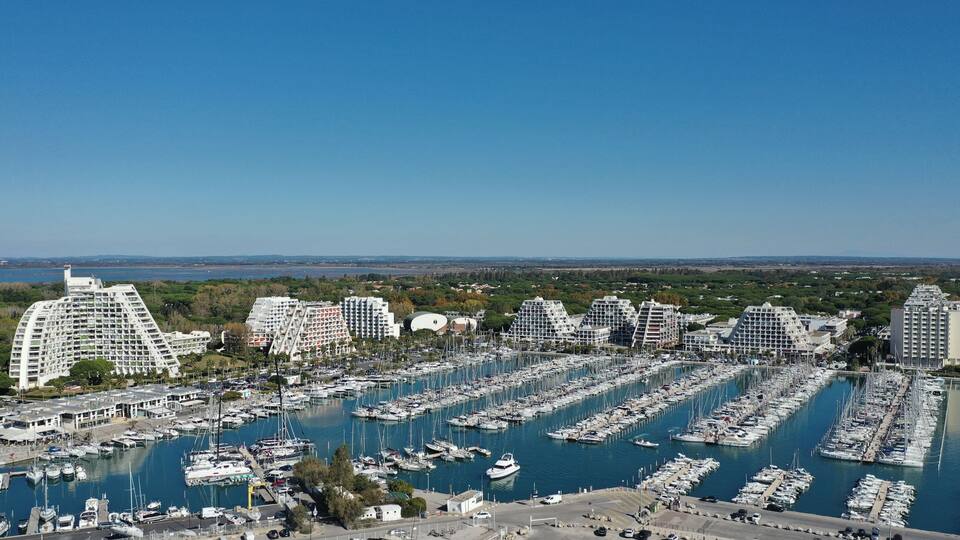 A marina with numerous boats docked, surrounded by modern buildings and a clear blue sky.