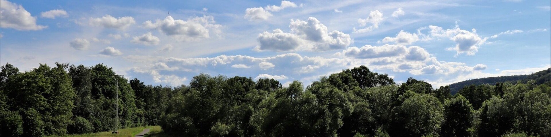 Un sendero sinuoso atraviesa un campo cubierto de hierba, con una colina boscosa al fondo y un cielo azul despejado con algunas nubes.