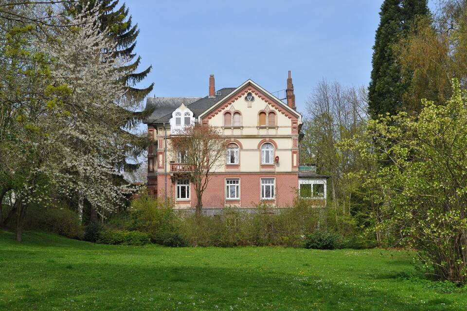 Un grand bâtiment historique à la façade en briques rouges et aux ornements blancs, entouré d’une végétation luxuriante et d’arbres en fleurs.