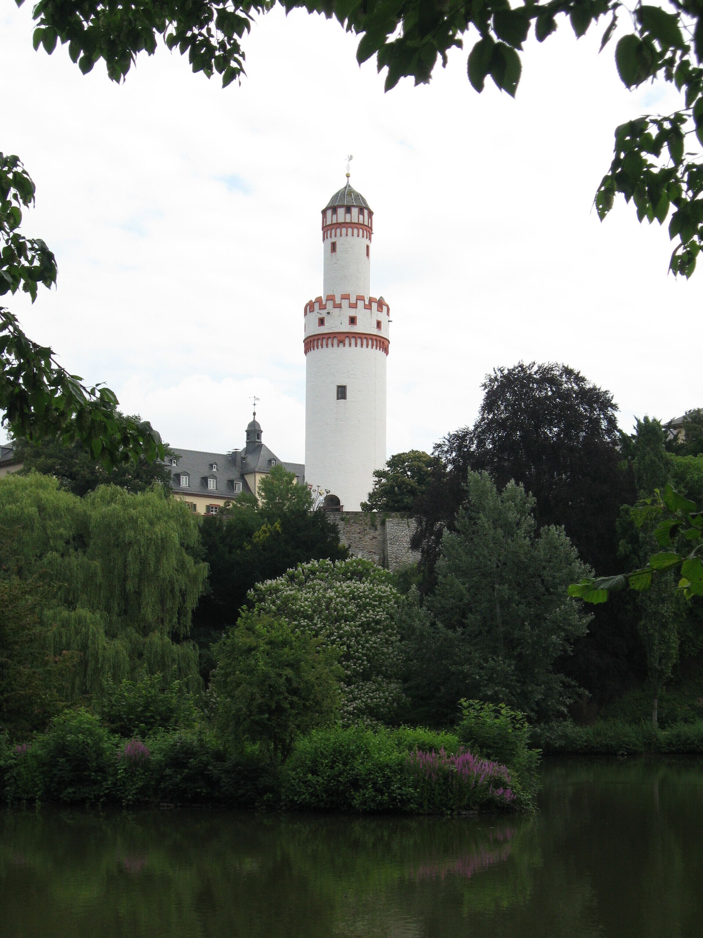 A white tower with a red band, surrounded by lush greenery and a calm body of water.