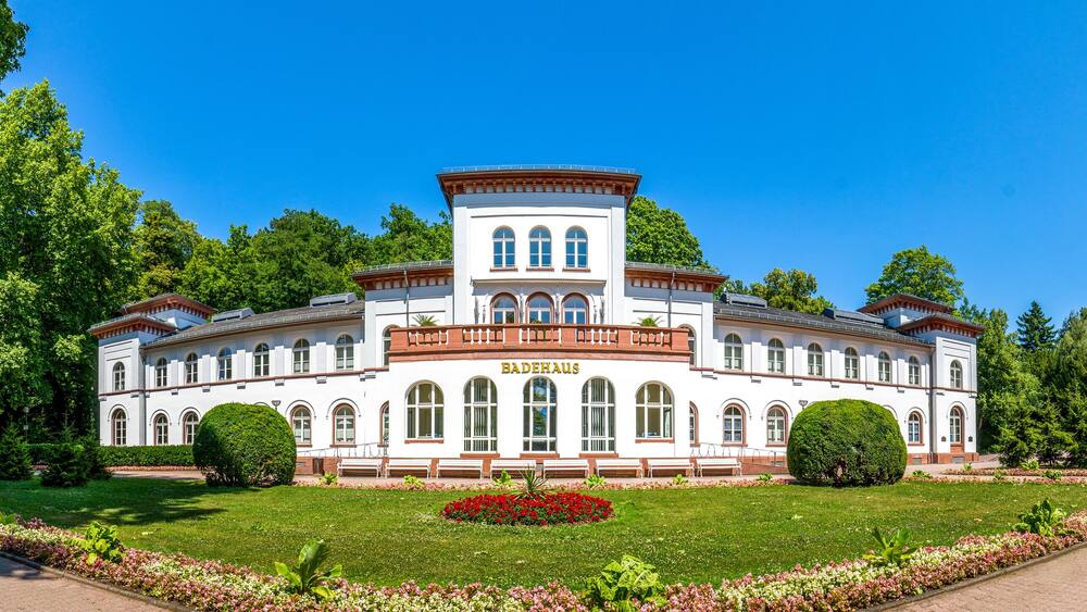 Un grand bâtiment blanc, doté de fenêtres en arc de cercle et d’une tour centrale, entouré d’arbres verdoyants et de jardins bien entretenus.