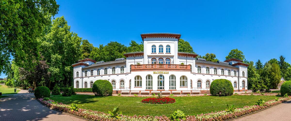 A large white building with arched windows and a central tower, surrounded by lush green trees and well-maintained gardens.