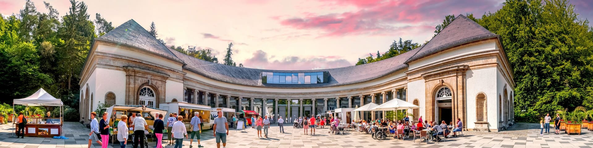 A spacious courtyard with a curved building, outdoor cafes, and a clear sky.