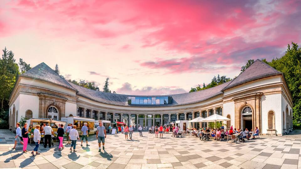 A spacious courtyard with a curved building, outdoor cafes, and a clear sky.