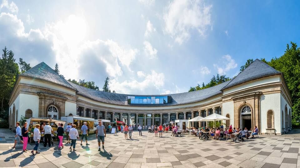 A spacious courtyard with a curved building, outdoor seating, and a clear sky.