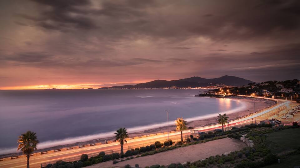 A coastal sunset with a beach, palm trees, and a cityscape in the distance.