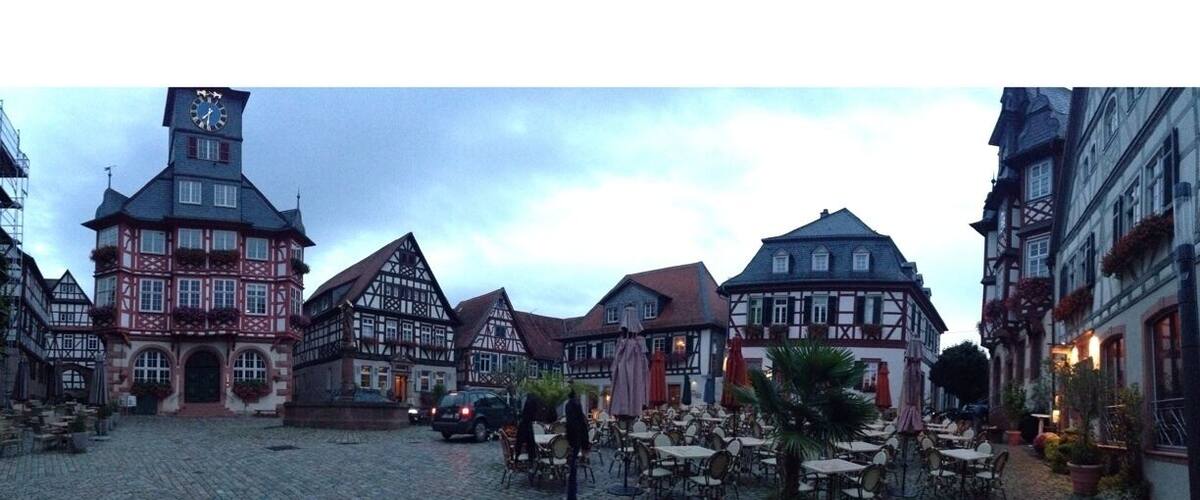 A cobblestone square with traditional half-timbered buildings, a clock tower, and outdoor seating areas.