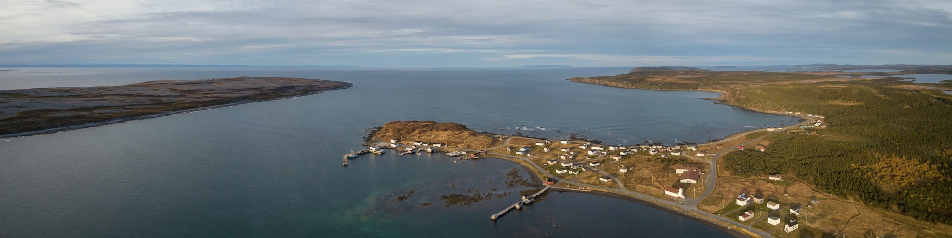 Vista aérea de un pueblo costero con un muelle, casas y un paisaje boscoso circundante.