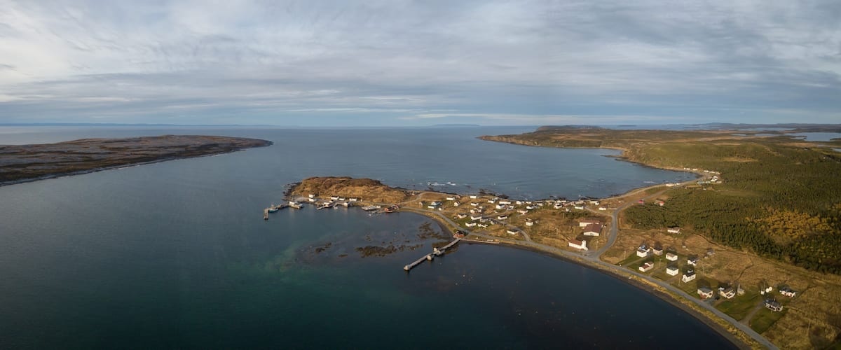 An aerial view of a coastal village with a pier, houses, and surrounding forested landscape.