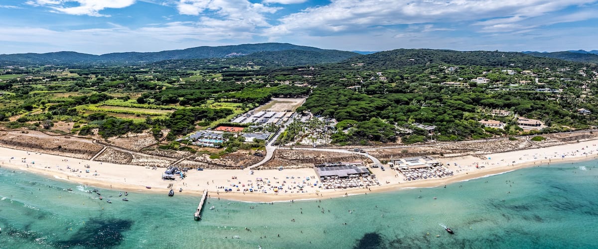 An aerial view of a coastal area with a beach, clear turquoise waters, and a backdrop of green hills.