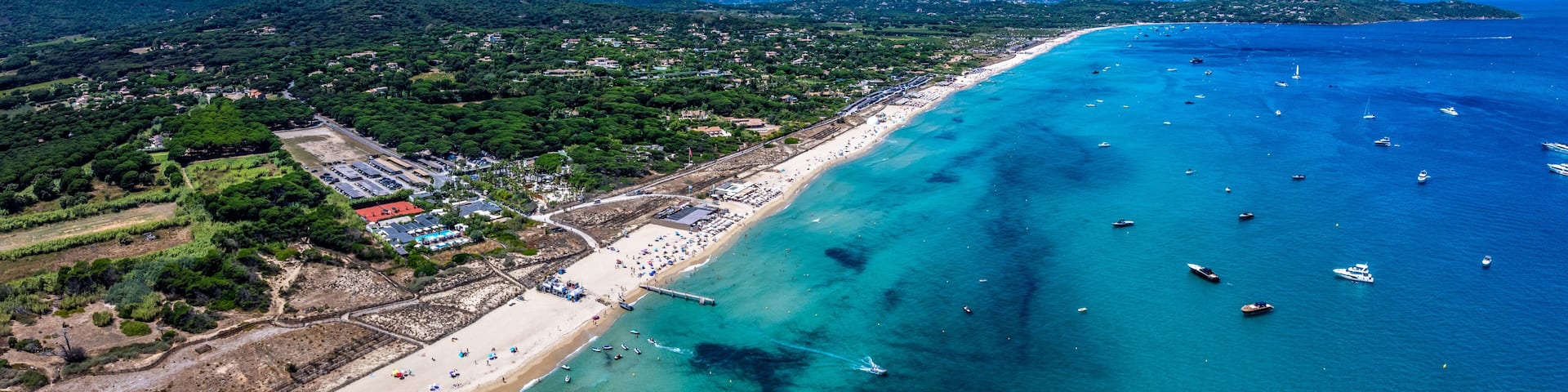aerial landscape view along Pampelonne Beach to Saint-Tropez, located at coastline of Ramatuelle, Côte d’Azur, France, with people on the beach in water, green wooded hilly landscape in background