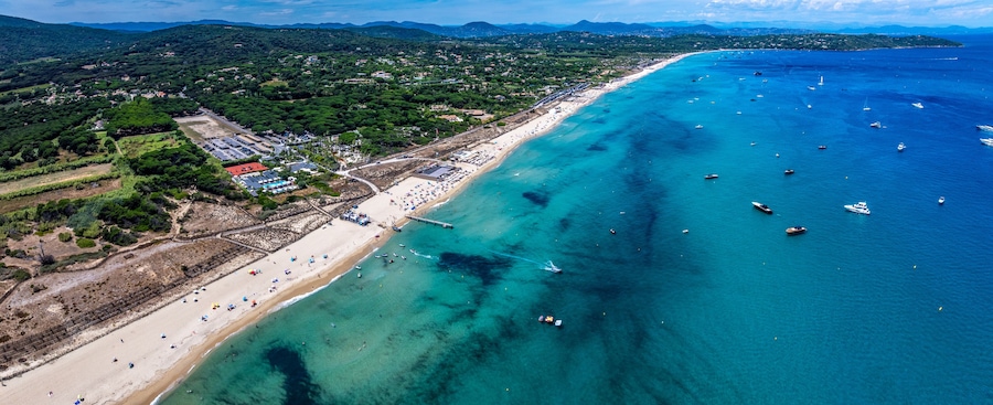 aerial landscape view along Pampelonne Beach to Saint-Tropez, located at coastline of Ramatuelle, Côte d’Azur, France, with people on the beach in water, green wooded hilly landscape in background