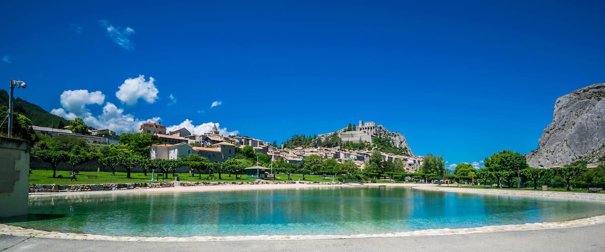 A coastal town with a clear blue sky, a large body of water, and a prominent hill with a castle-like structure on top.