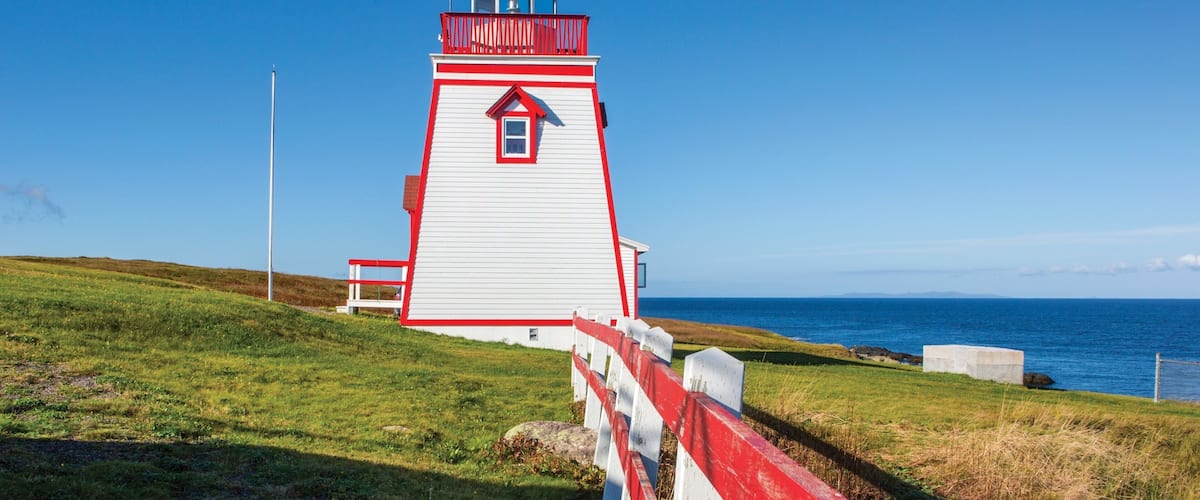 A white and red lighthouse with a balcony, situated on a grassy hill by the sea.