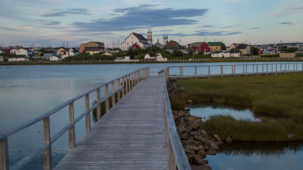 A wooden pier extending into calm water, surrounded by grassy banks and a row of houses with a church in the background.