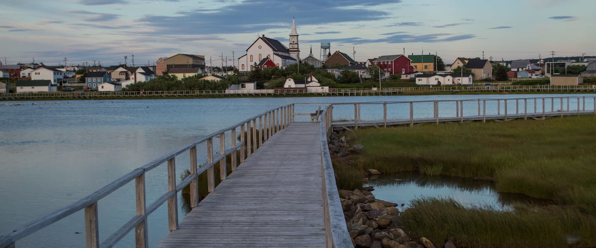 A wooden pier extending into calm water, surrounded by grassy banks and a row of houses with a church in the background.