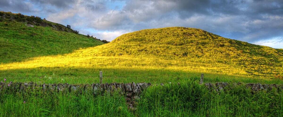 A grassy hill with a stone wall in the foreground and a cloudy sky.