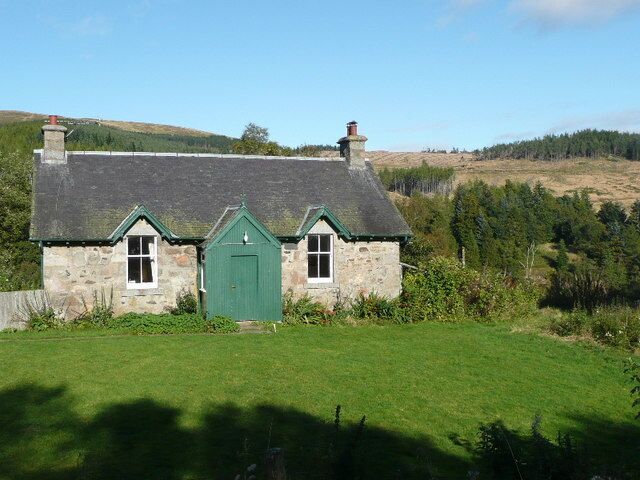 A stone house with a green door and a thatched roof, surrounded by a grassy field and trees.