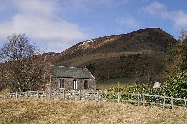 A stone church with a slate roof, surrounded by a stone fence, set against a backdrop of rolling hills and a clear sky.