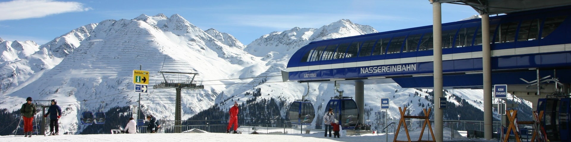 Un centro de esquí con un telesilla, montañas cubiertas de nieve y un edificio azul con la leyenda "Nassereinbahn".