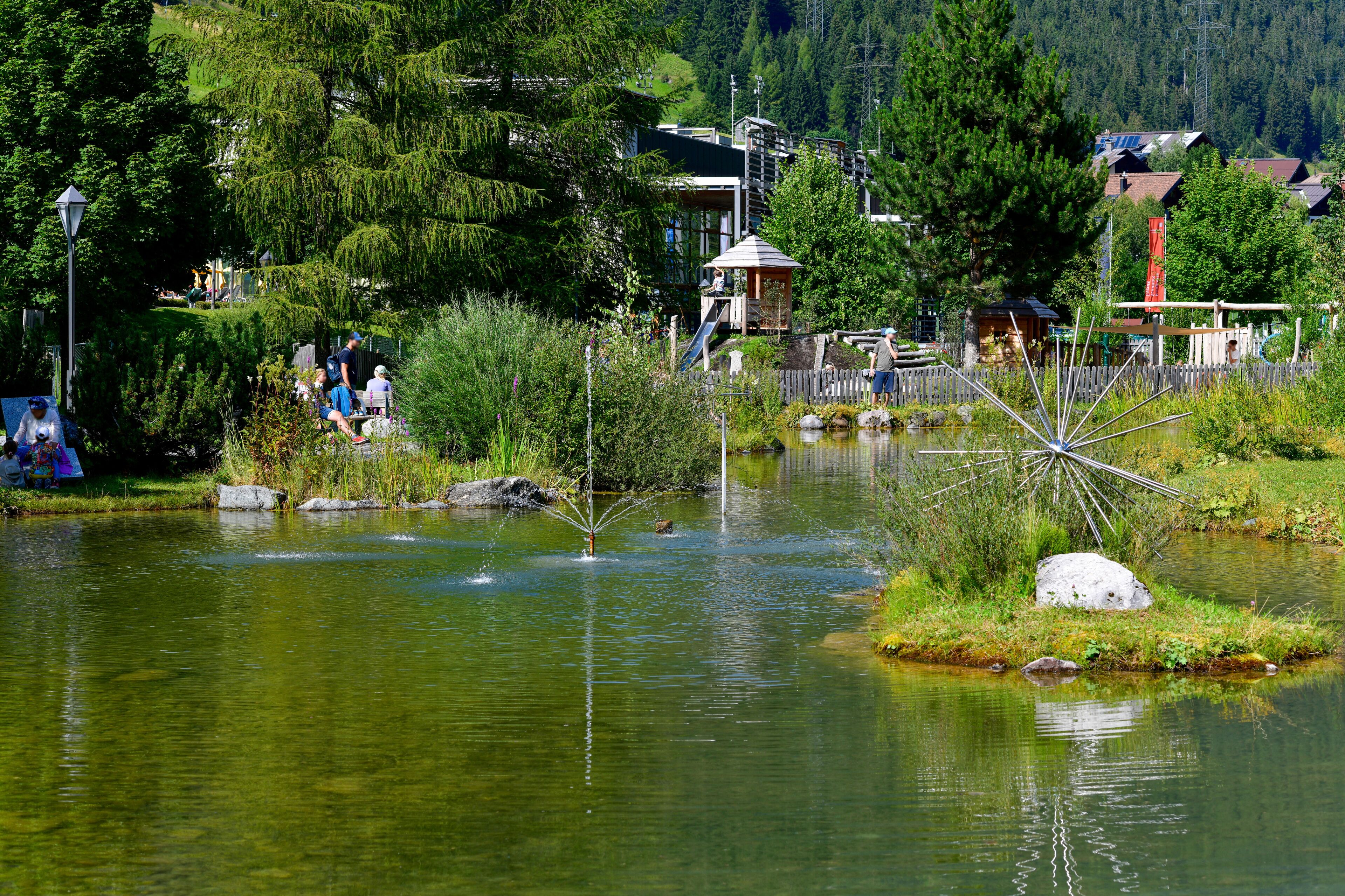 A tranquil pond with a fountain, surrounded by lush greenery and a wooden fence.
