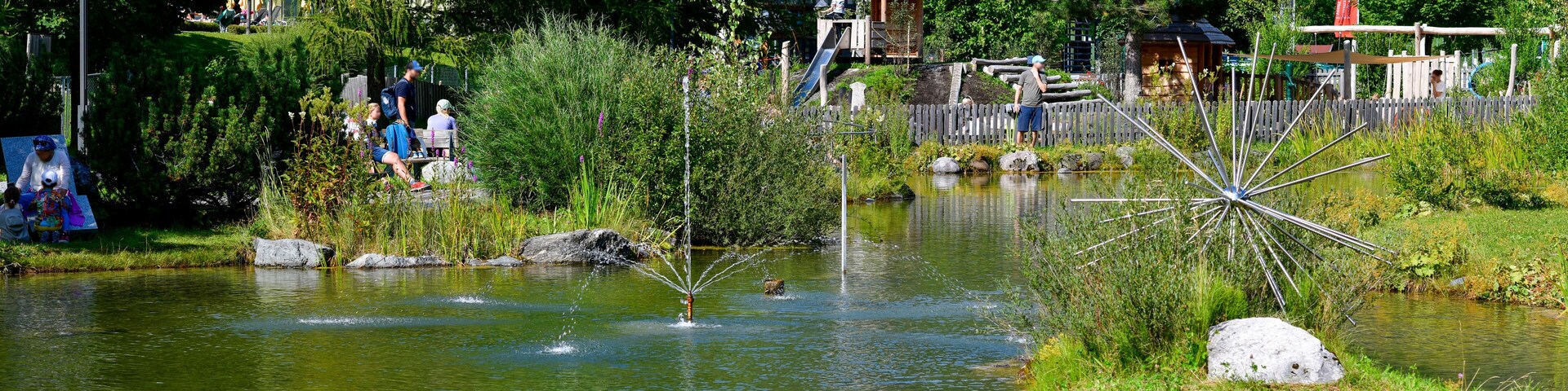 A tranquil pond with a fountain, surrounded by lush greenery and a wooden fence.