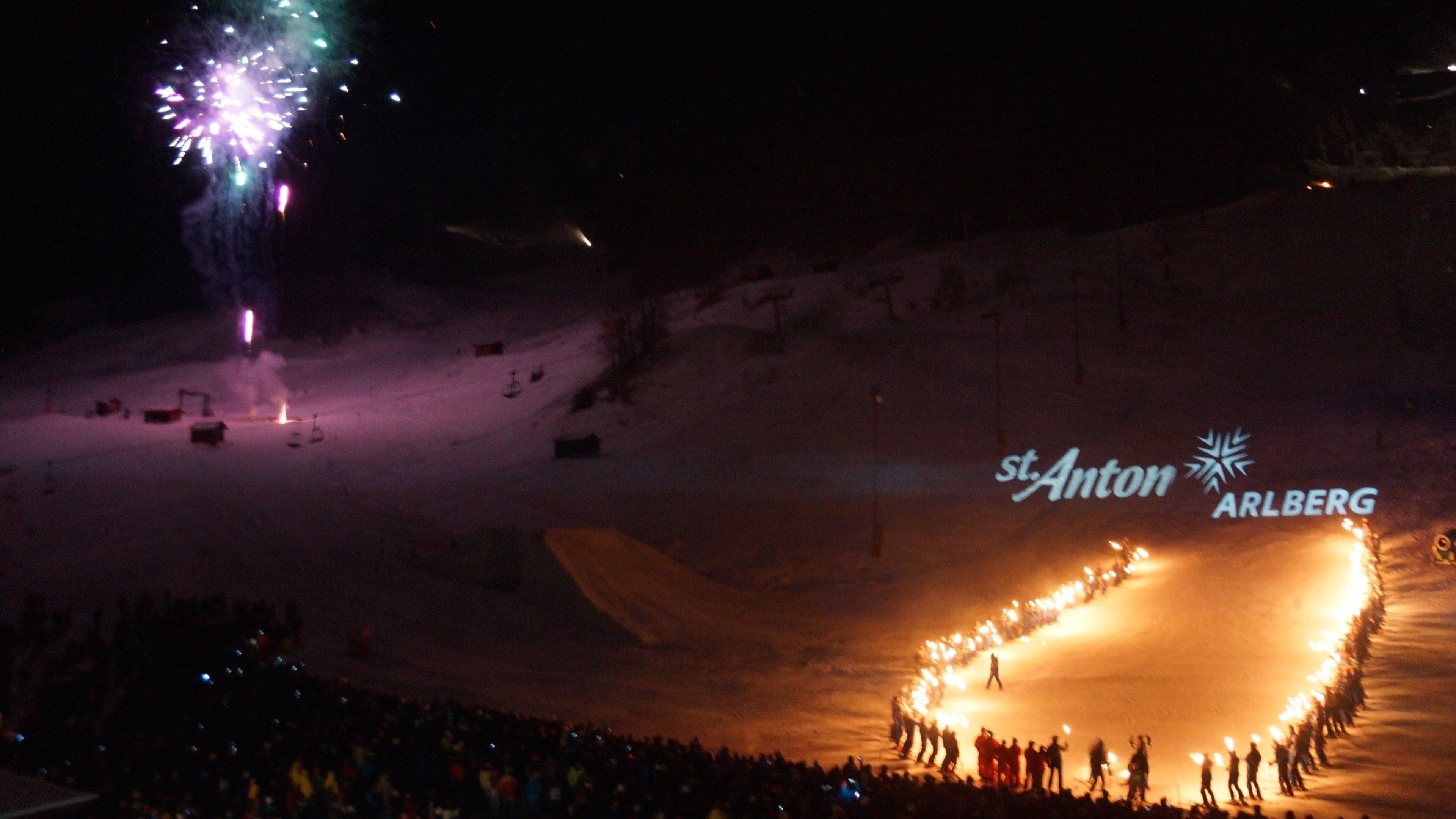 A snow-covered ski slope with a crowd forming a heart shape, and the St. Anton ARLBERG logo visible.
