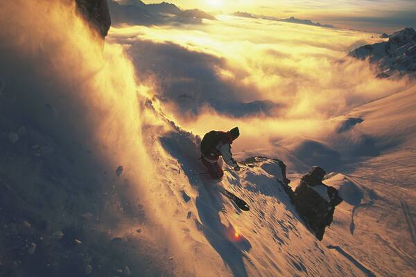 Ein Skifahrer fährt einen schneebedeckten Berghang hinab, darunter Wolken.