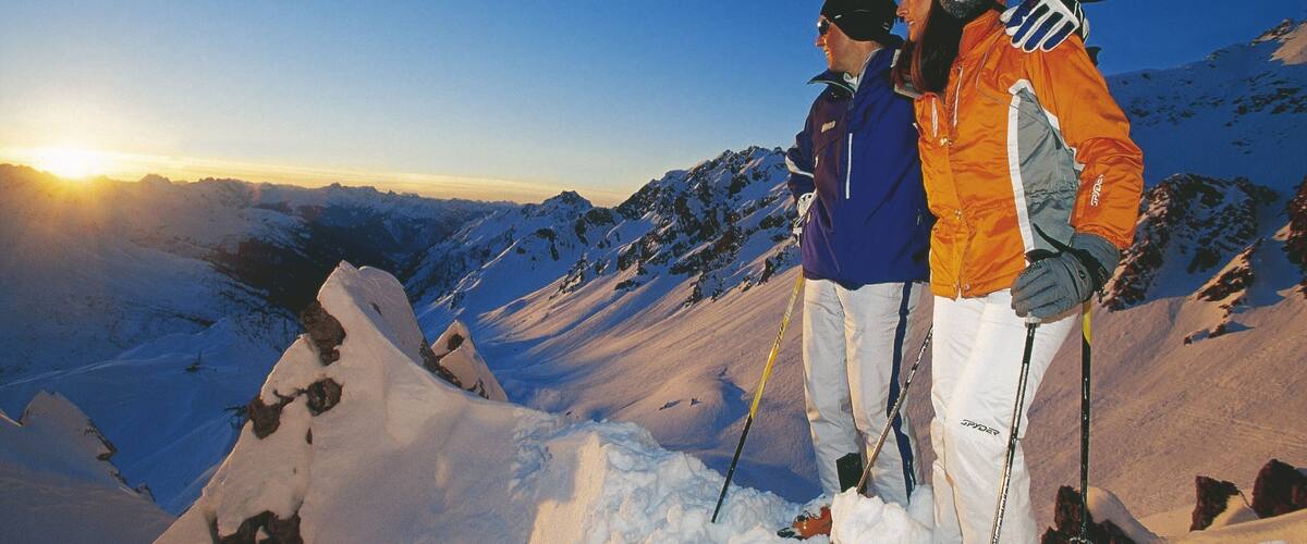 Two skiers on a snowy mountain peak with a clear sky and the sun setting in the background.
