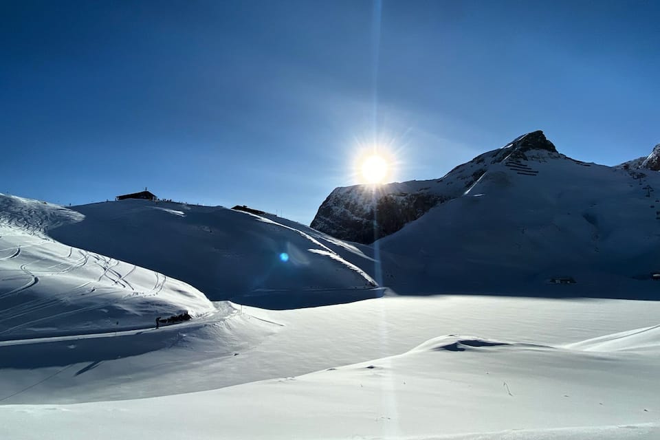 A snowy mountain landscape with a clear sky and the sun shining through.
