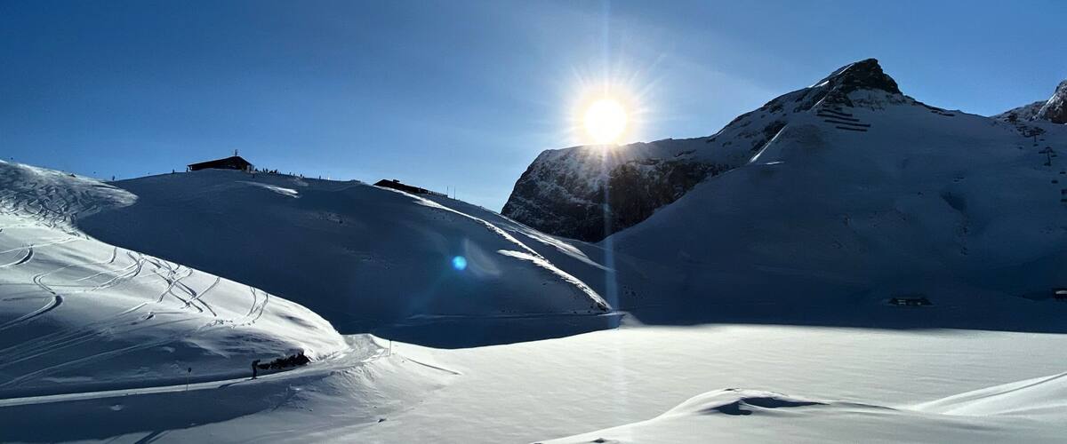 A snowy mountain landscape with a clear sky and the sun shining through.