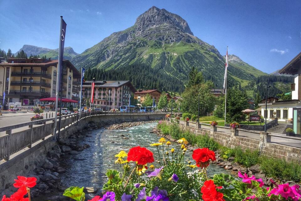 Um rio serpenteando por uma cidade, com flores coloridas em primeiro plano, uma montanha ao fundo e edifícios ao longo da margem.